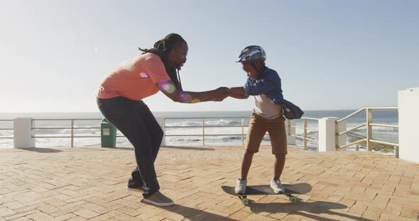 Video of happy african american father learning son how to skateboard on promenade