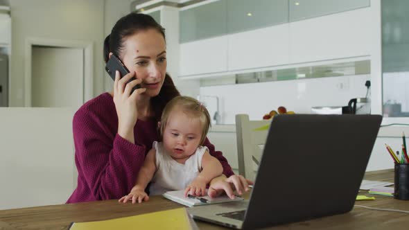 Caucasian mother holding her baby talking on smartphone and using laptop while working from home