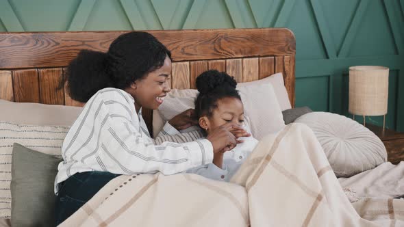 African Mother Babysitter Older Sister with Little Girl Child Daughter Lie on Bed at Home Looking at