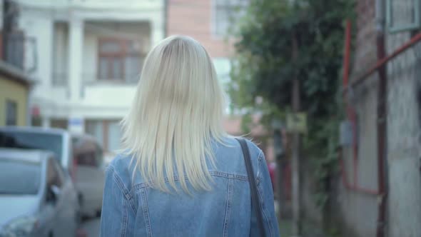 Rear view of young stylish blonde in denim jacket walking through city center