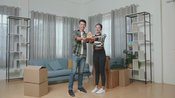 Couple With Cardboard Boxes On The Floor Smiling And Showing House Model To Camera In The New House