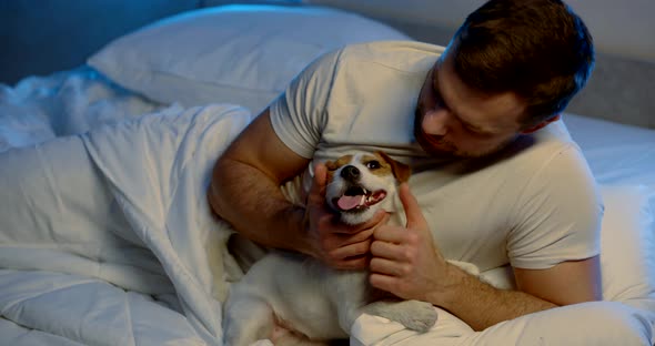 Male Owner Is Inspecting Teeth of His Jack Russell Puppy, Lying in Bed at Night