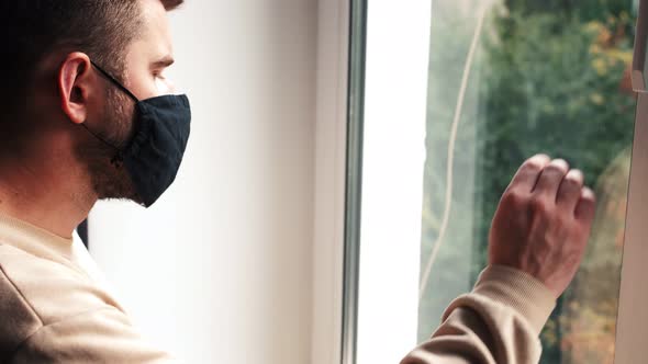 A Sad Young Man Is Sitting at Home in Quarantine. A Man Looks Out the Window and Puts His Hand To