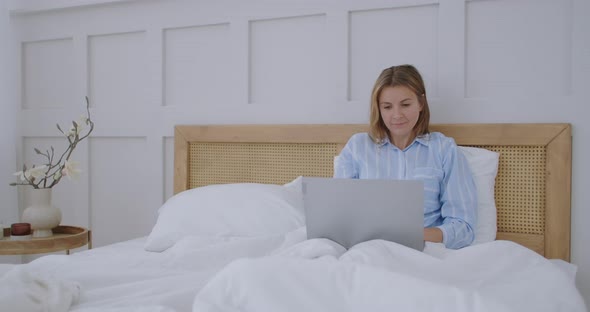 Happy Woman with Laptop Lying in Bed and Smiling. Girl in a Man's Shirt Working on a Laptop in the