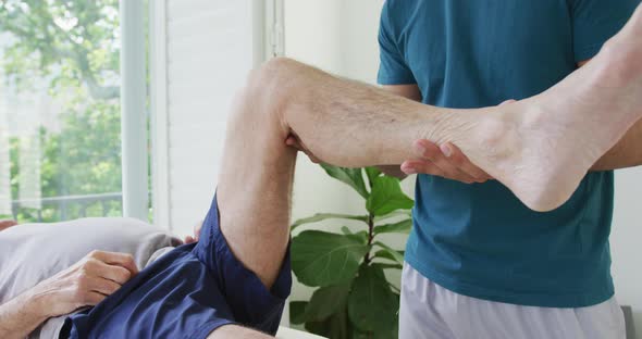 Biracial male therapist examining while lifting senior man's leg at health club