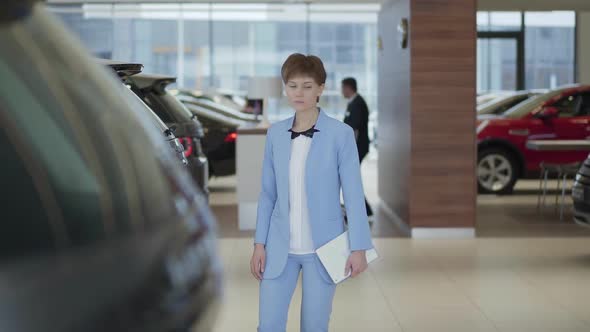 Young Pretty Woman with Tablet in Hands in Formal Wear Blue Suit Walking Between Rows of New Cars