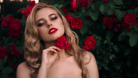 Fabulous Girl with Red Lips in Red Dress on Awesome Summer Roses Background. Fantasy Woman Portrait