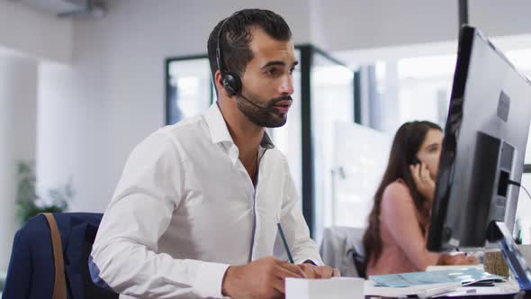 Mixed race businessman sitting at desk talking using phone headset and computer in busy office