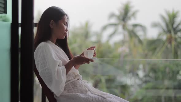 Asian lady, wearing white bathrobe , stirring a hot morning coffee and taking a sip while sitting on