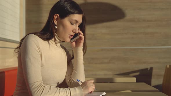 Young Woman in a Cafe Talking on the Phone and Making Notes in a Notebook