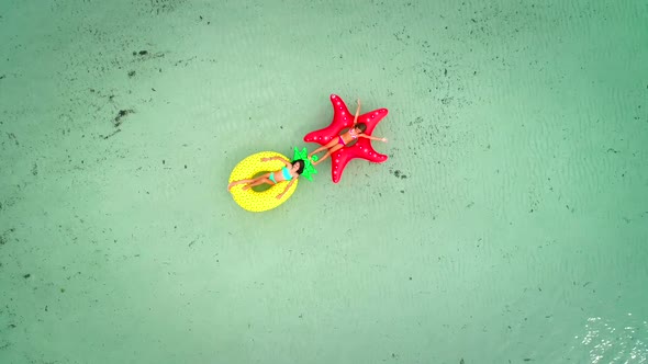 Aerial view of two girls floating on inflatable mattress in transparent sea, holding each other.