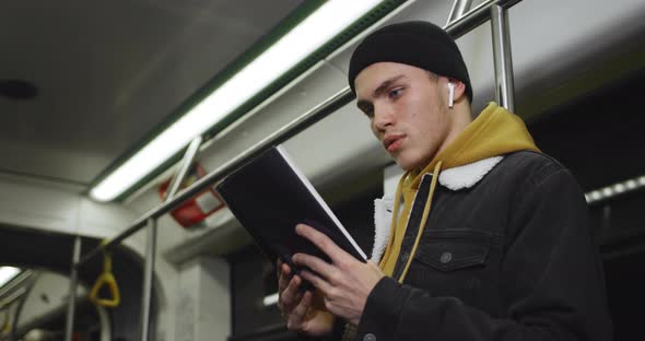 Side View of Young Man with Wireless Earpods Turning Pages While Enjoing Story. Millennial Handsome