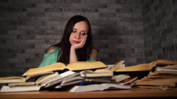 Tired Female Student Reading Among Books. Pensive Young Woman Sitting at Table with Pile of Book and