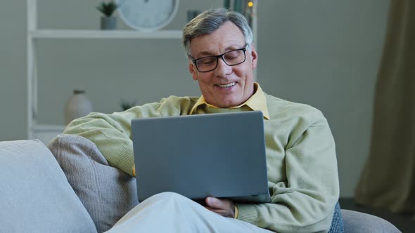 Happy Grandfather Sit on Sofa Living Room Mature Elderly Caucasian Man Looking at Screen Laptop Work