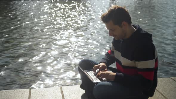 Handsome young man sitting on the banks of the canal in Amsterdam typing a text on the notebook. Net