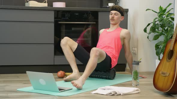 Young fit guy with fitness roll doing rehab exercises using online training app on notebook at home