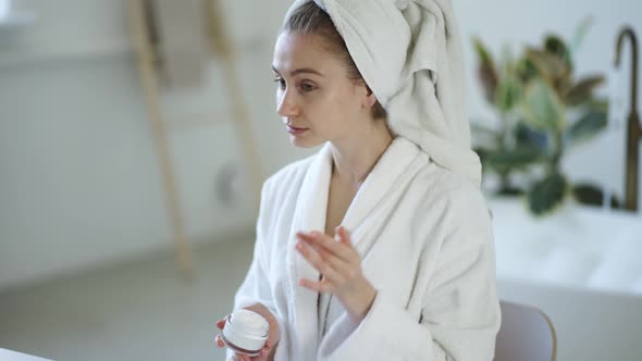 Young Woman in White Bathrobe and with Towel on Head Applies Moisturizer Cream on Her Face