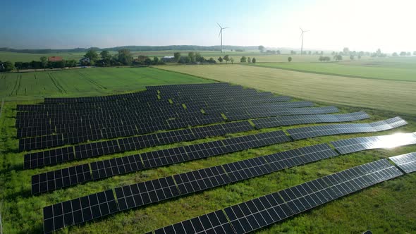 Aerial view showing solar panel farm and rotating wind turbine in background on green rural farm fie