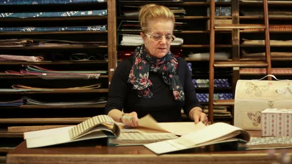 Senior Woman in Her Paper Shop is Selecting Pieces of Paper in Front of Wooden Shelf Where are