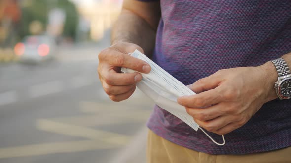 A Man Stands By the Road or at a Bus Station Waiting for Transport and Holds a Medical Mask in His