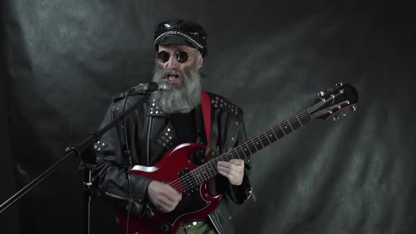 Bearded rock musician in leather jacket and cap plays red electric guitar and sings on stage