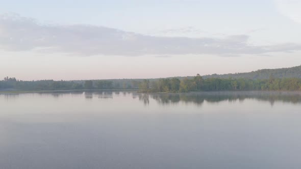 Aerial flying over calm lake while fog sets in during golden hour