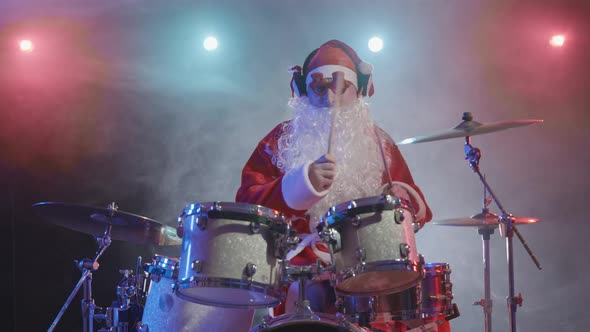 Santa Claus in a Red Costume with Beard and Funny Glasses Plays the Drums in a Dark Studio with