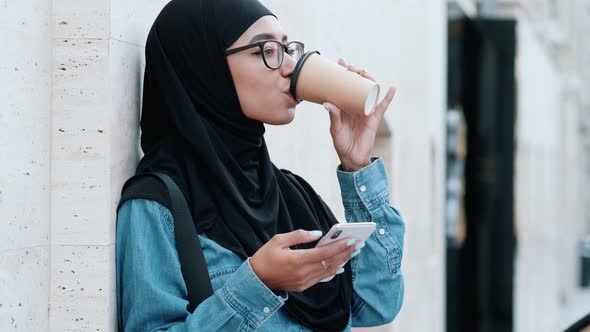 A pleased arabian woman wearing national hijab is using her phone while drinking coffee