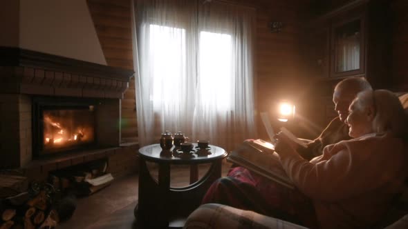 An Elderly Couple Considers a Family Photo Album Wrapped in a Blanket Sitting By a Cozy Fireplace