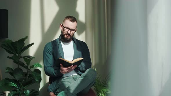 Bearded Young Man Sitting Near Window and Reading a Book at the Sunrise