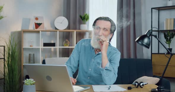 Old Man with Wrinkled Face Sitting in front of Camera and Smoking a Cigar with Pleasure