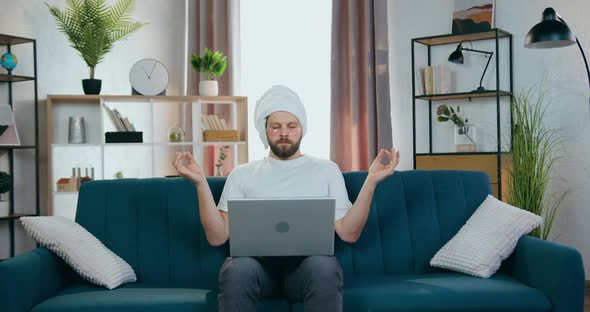 Guy with Patches Under Eyes and Shower Towel Around His Head Sitting at Home with Laptop