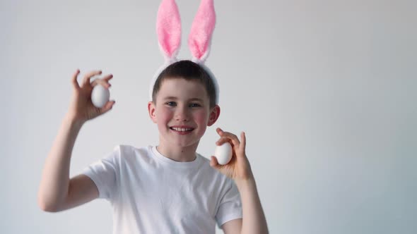 Playful boy with bunny ears keeping white eggs near eyes on white background. Happy child