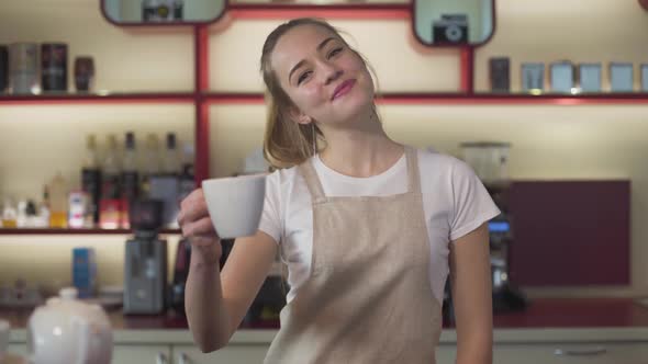 Smiling Young Caucasian Woman Showing Coffee Cup at Camera and Smiling. Professional Female Barista
