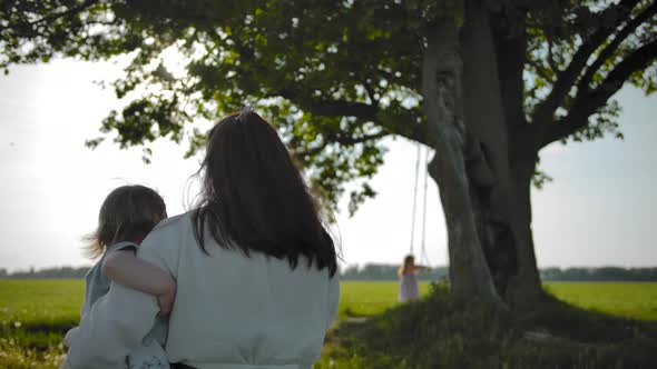 Mom in a White Dress with Daughter in Her Arms Goes To an Old Oak Tree.