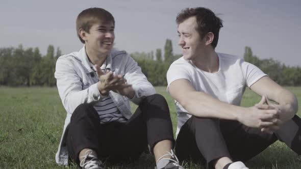 Two Young Positive Men Sitting on Green Summer Meadow Laughing