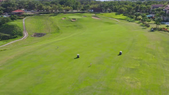 Aerial drone view of two golf cars and two people who walk in a golf course with a luxury resort in