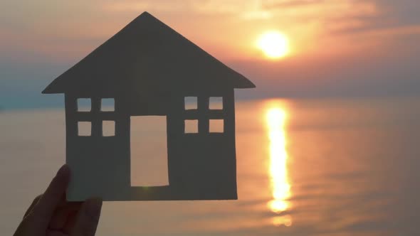 Hand Holding Cut Paper House Against of Sea at Sunset