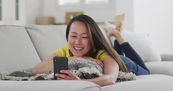 Happy asian woman lying on sofa, having video call on smartphone at home