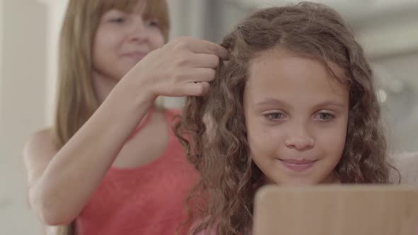 Beautiful Young Mother Braids Her Cute Daughter Looking in the Mirror. Family Relationships