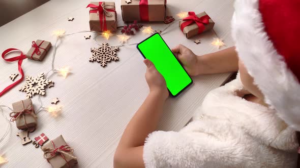 Cute girl in Santa hat holding smartphone with vertical green screen.