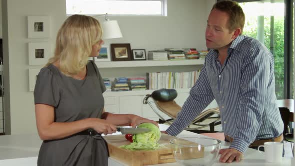 Female preparing salad in kitchen, male talking