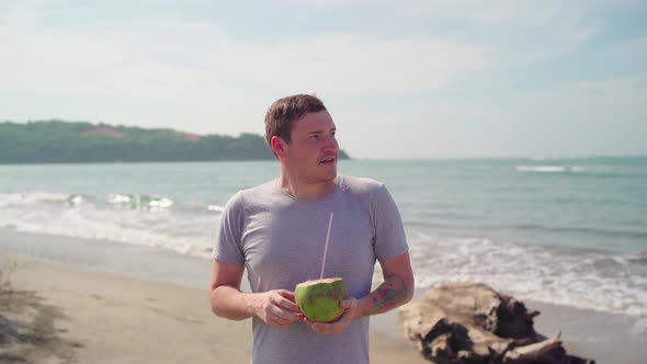 Relaxed adult guy standing and drinking coconut on sandy beach. 