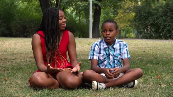 A Young Black Mother and Her Son Sit on Grass in a Park and Talk About Something