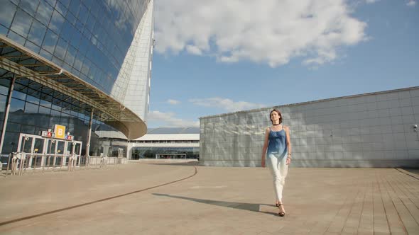 Pretty Young Girl Walking In Highheels In A Long Light Dress Along The Huge Glass Building 