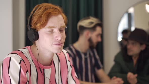 Relaxed Redhead Man Listening to Music in Headphones with Blurred Friends Talking at Background