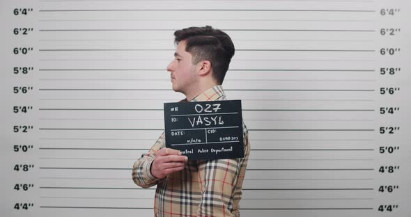 Portrait of Male Criminal in Shirt Turning to Sides While Holding Sign for Photo in Police Station