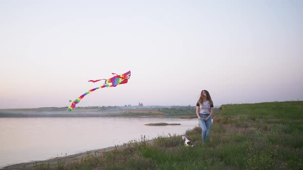 Young Happy Woman and Het Little Dog Walking with Flying Kite on a Glade at Sunset Slow Motion