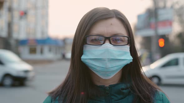 A Young Girl in Glasses and a Medical Mask Stands on the Street