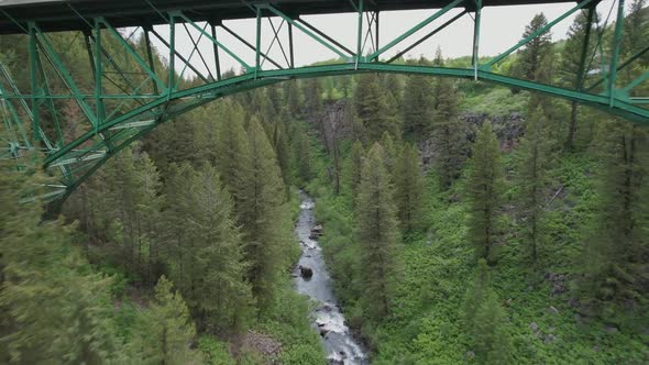 Drone flies under a green painted steel bridge.  The bridge runs over a creek surrounded by pine tre